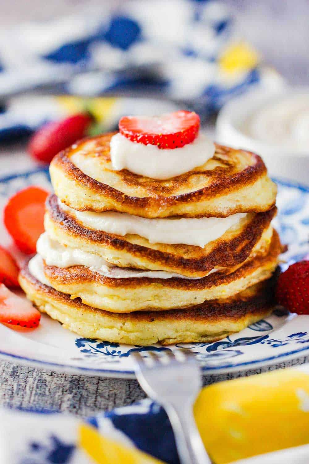 A stack of lemon ricotta pancakes with vanilla sauce on a antique plate with a fork nearby and strawberries.