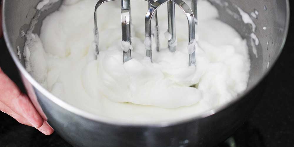 A close-up view of a hand mixer making stiff peaks from egg whites in a metal bowl.