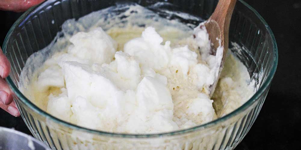 A wooden spoon folding stiff peak eggs whites into a pancake batter in a large glass bowl.
