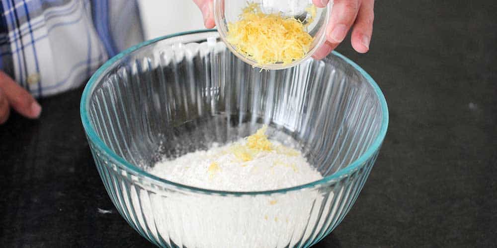 A hand dropping a small bowl of lemon zest into a glass bowl of a flour mixture.