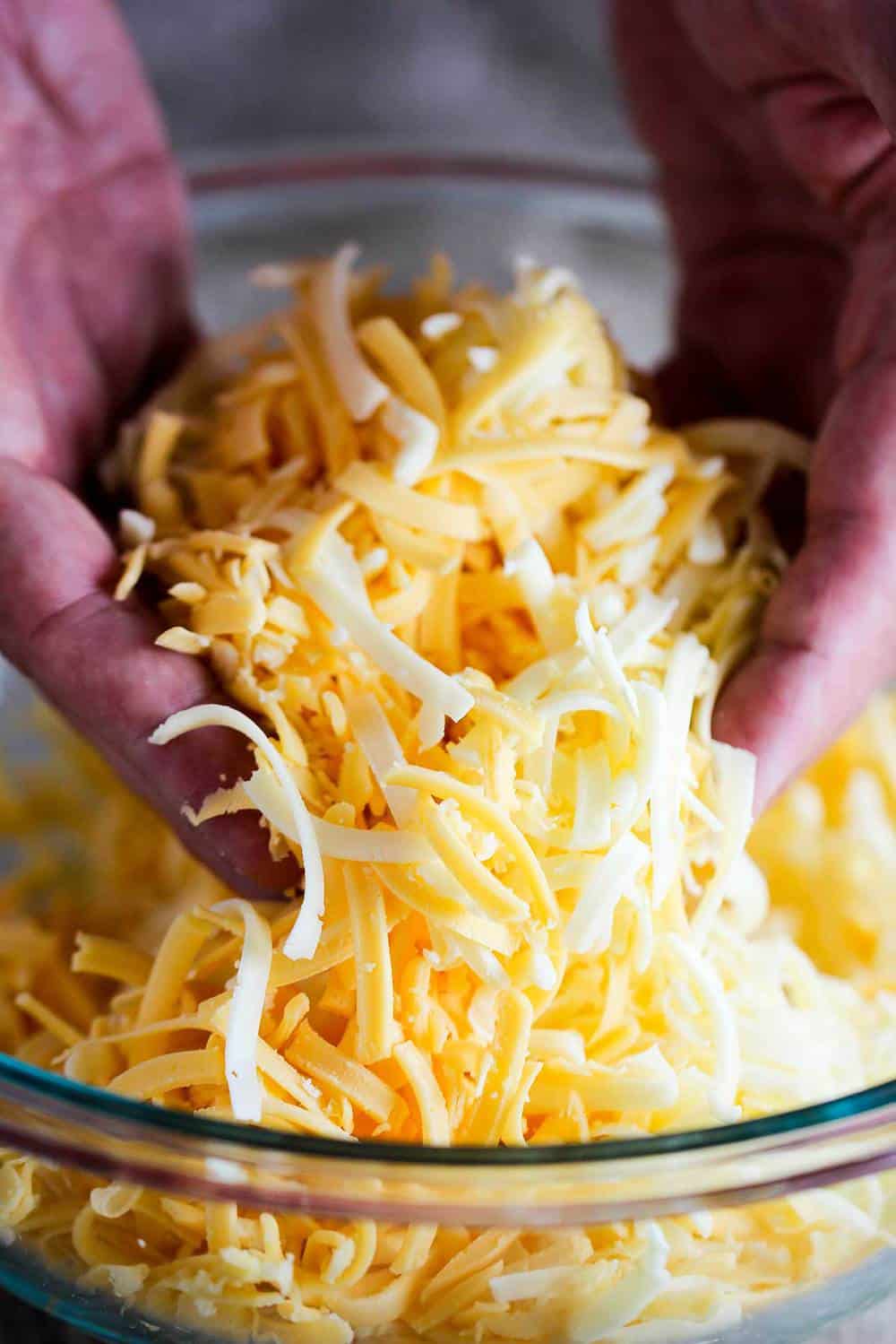 Shredded yellow and white American cheese being lifted out of a glass bowl.