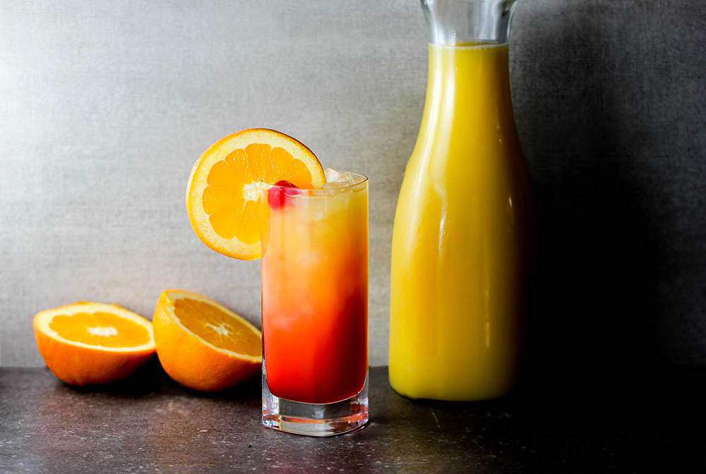 Classic Tequila Sunrise with oranges and fresh orange juice in the background.