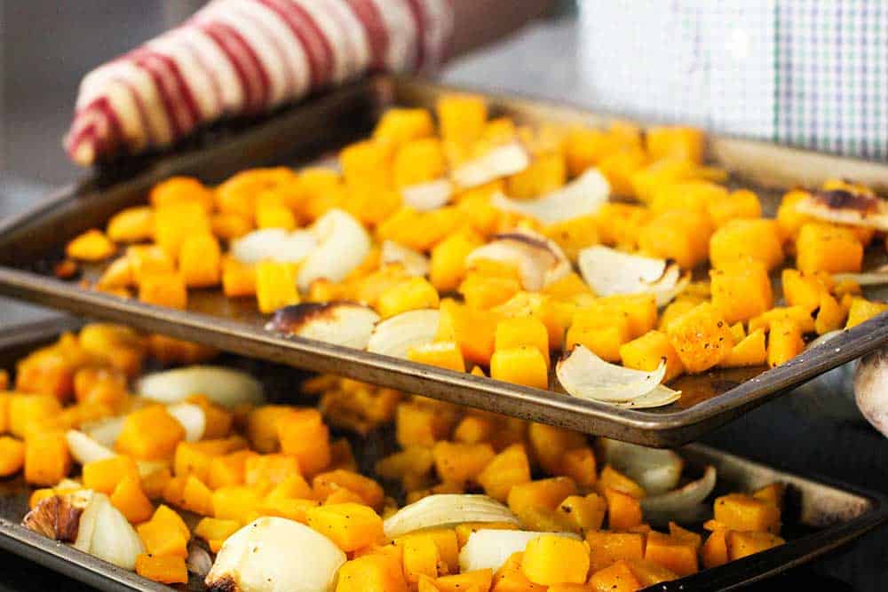 Two hands with oven mitts holding a baking sheet with roasted butternut squash and onion on it.