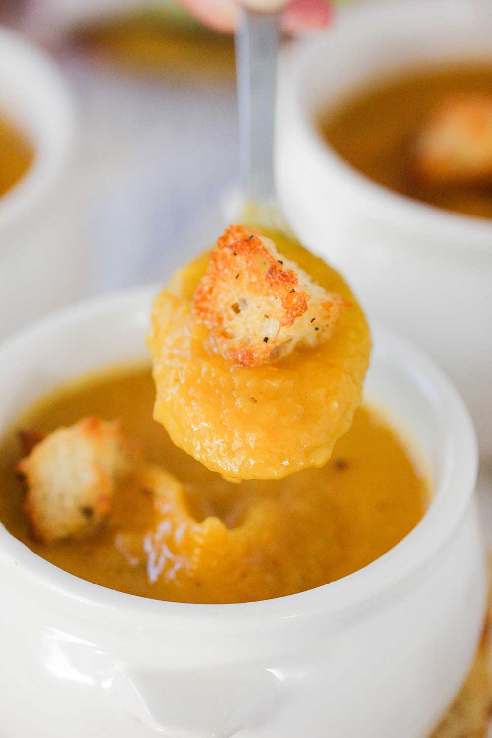 A hand holding a spoon full of roasted butternut squash soup over a small crock of soup with croutons on it.