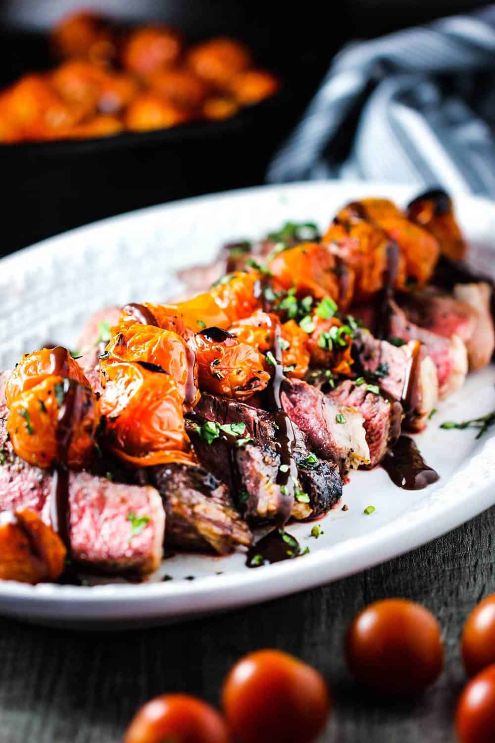 A white platter of seared ribeye with blistered tomatoes and bordelaise sauce on top