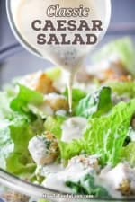 Caesar salad dressing being poured from a small glass jar onto a bowl of torn lettuce and homemade croutons.