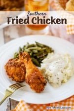 A white dinner plate filled with pieces of Southern fried chicken, a pile of mashed potatoes with cream gravy, and a helping of green beans.