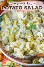 A colorful serving bowl filled with homemade best-ever potato salad with fresh dill and a wooden spoon inserted into the side of the salad.