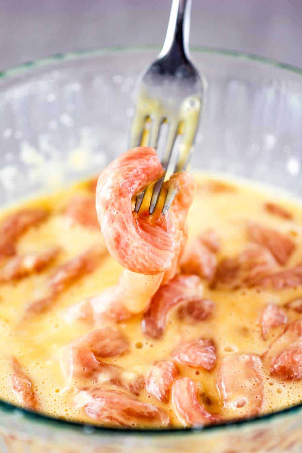 A glass bowl of uncooked pork strips in an egg.