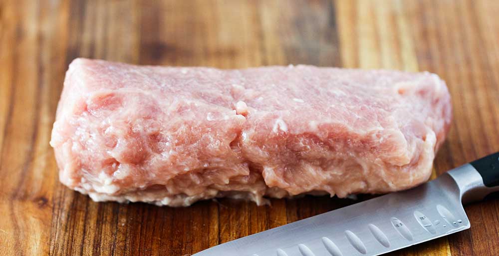 An uncooked pork tenderloin on a cutting board with a chef's knife next to it.