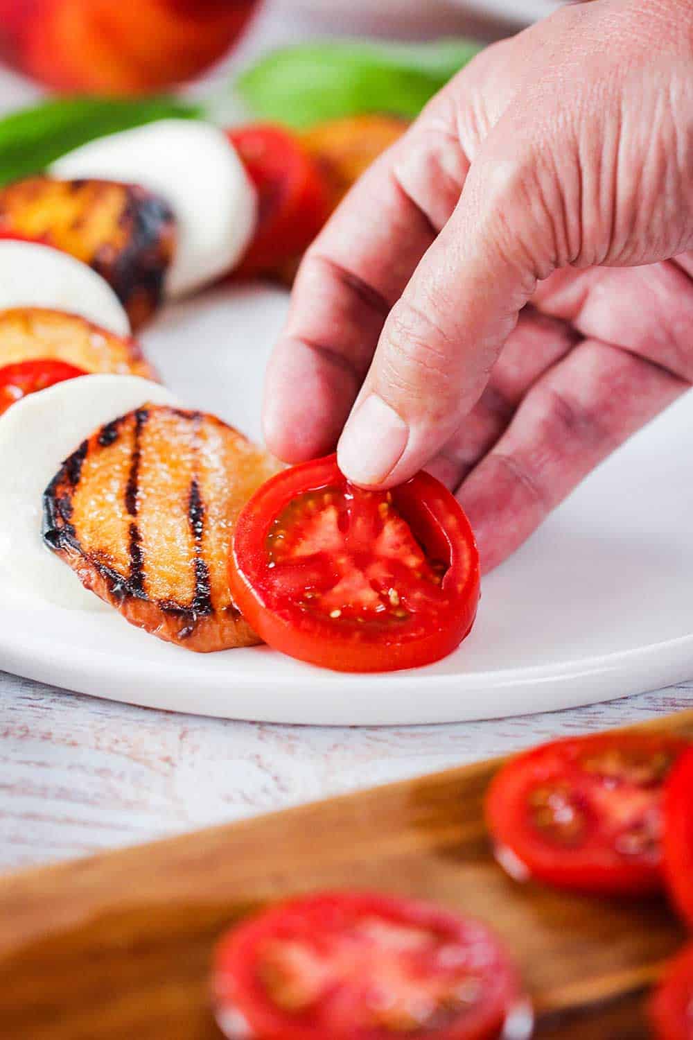 A hand layering a tomato for caprese salad with grilled peaches on a white plate.
