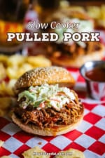 A straight-on view of a slow cooker pulled pork sandwich sitting on a red checkered sandwich wrapper with kettle chips scattered around it.