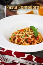 A large white bowl filled with pasta pomodoro next to a red-checkered napkin and a glass of red wine.