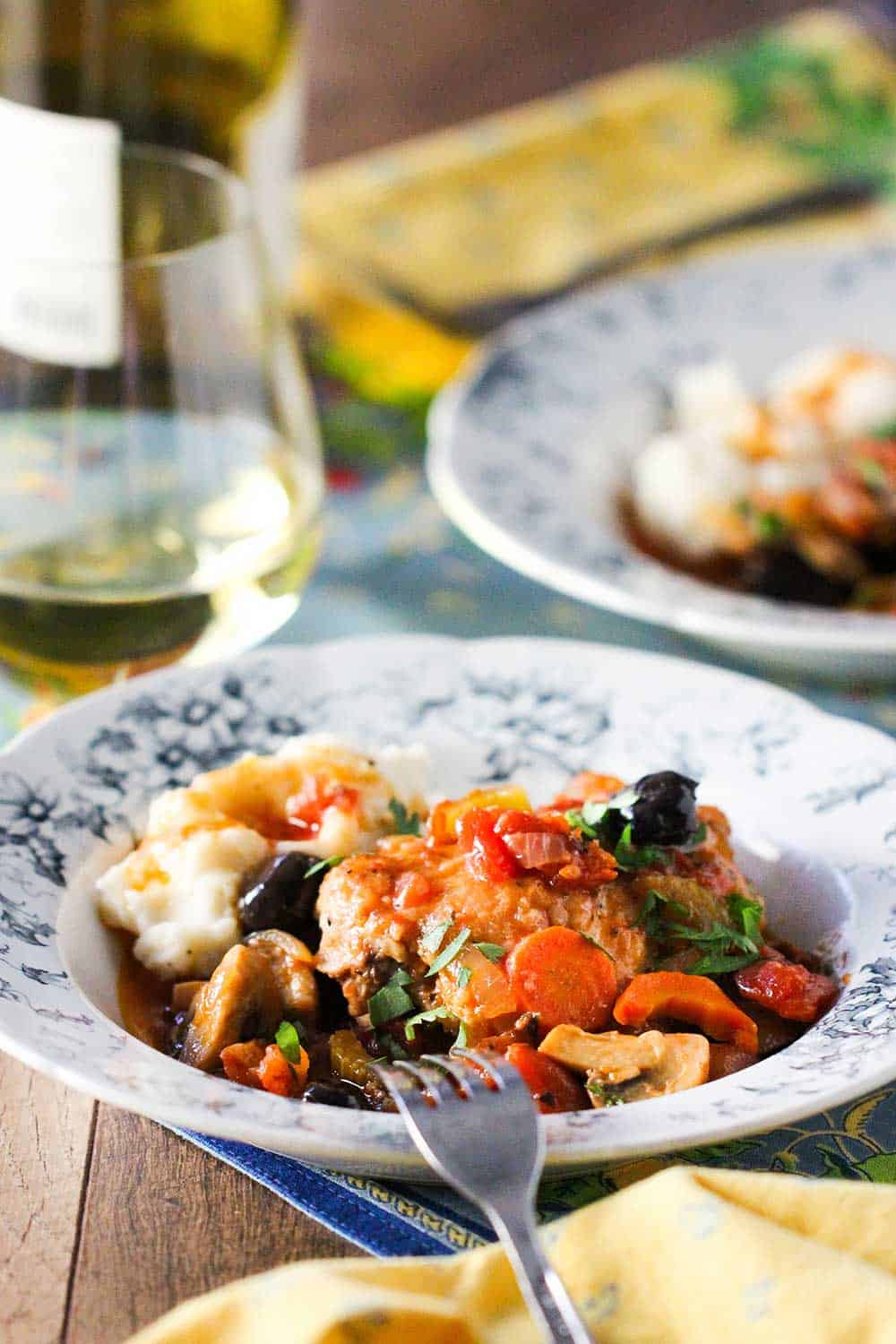A French dish filled with seared chicken with a tomato, mushrooms and vegetables with a bottle of white wine next to it.