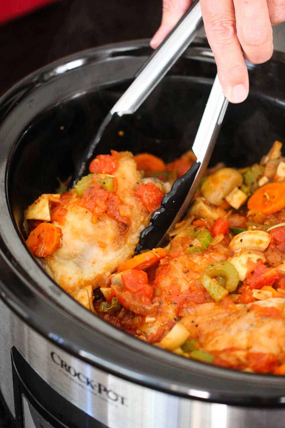 A pair of tongs nestling seared chicken thighs into a stew sauce in a crock-pot for Provençal chicken stew.