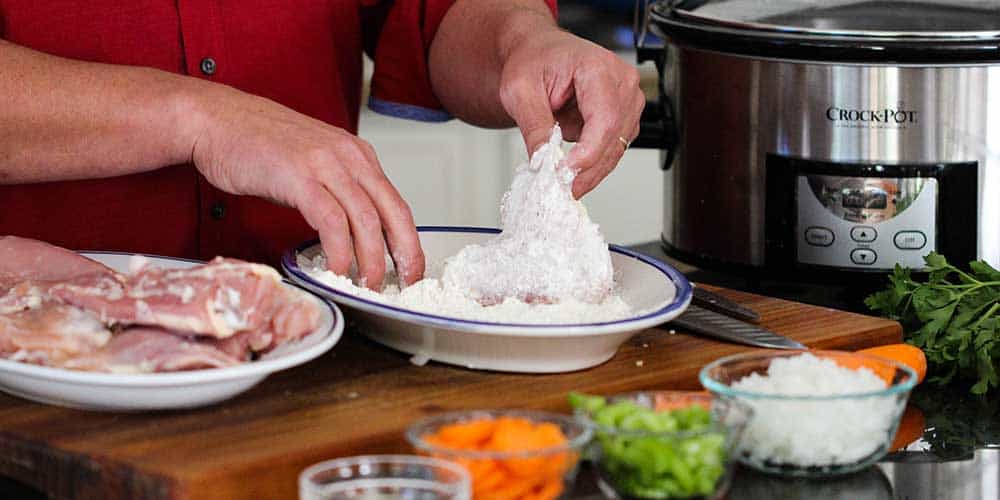 Two hands dredging skinless chicken thighs in a flour mixture for Provençal chicken stew.