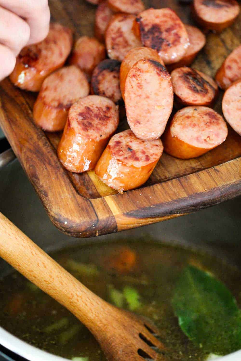 A wooden cutting board with sliced seared smoked sausage that is being transferred into an Instant Pot with chicken stock and bay leaves.