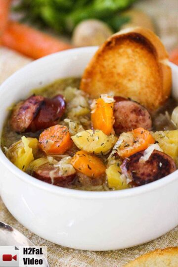 A large white bowl filled with smoked sausage stew and a piece of toasted bread stuck in the side with carrots in the background.