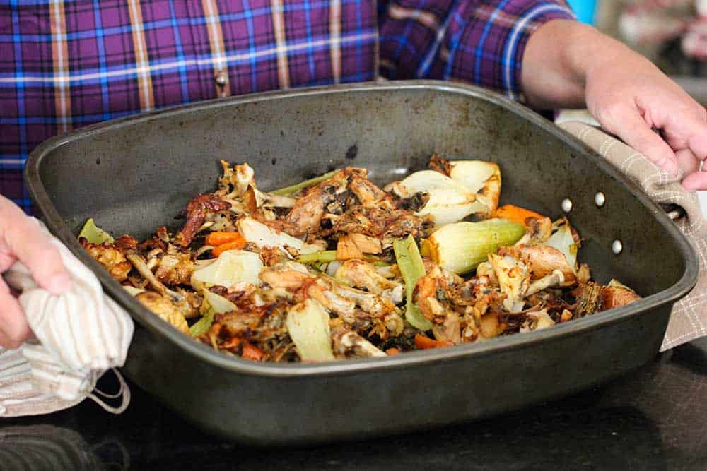 Two hands holding a large roasting pan of roasted chicken bones and vegetables.