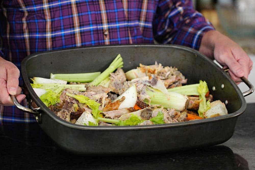 Two hands holding a roasting pan filled with chicken bones and vegetables.