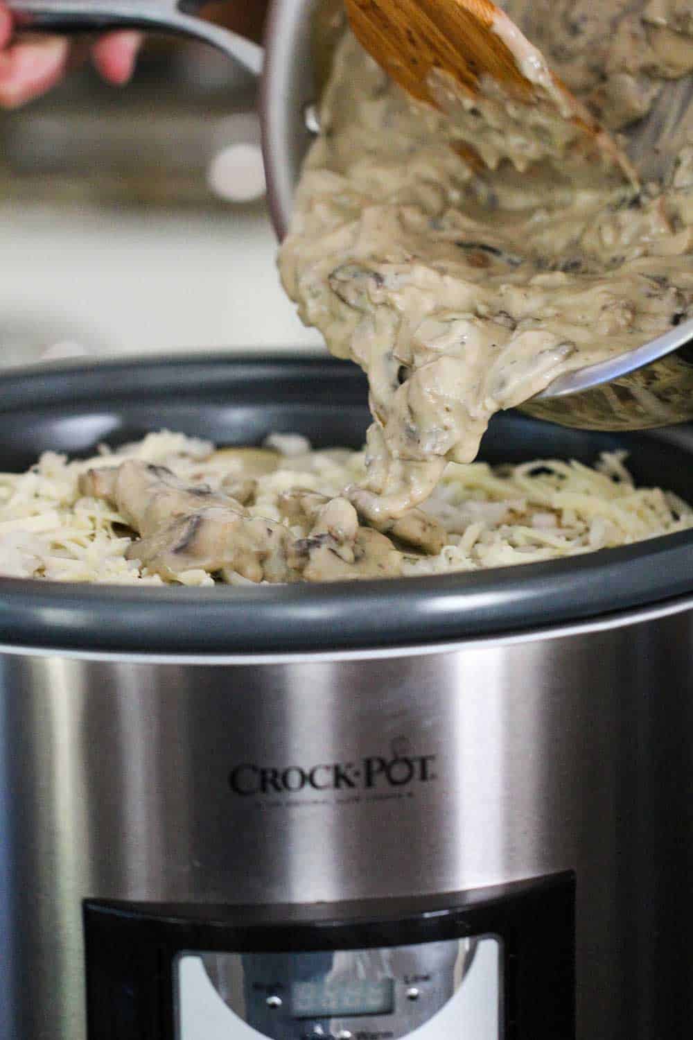 A wooden spoon transferring a mushroom sauce into a slow-cooker filled with potatoes, ham and cheese.