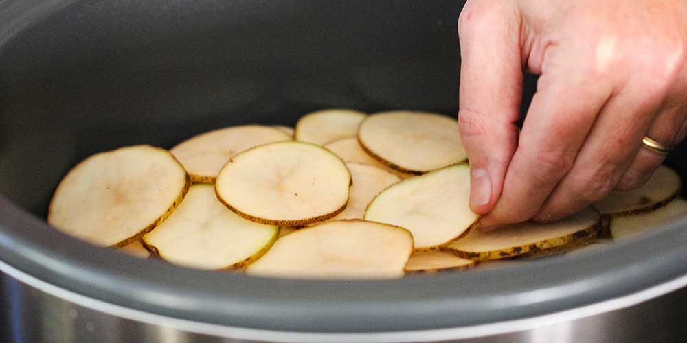 A hand adding a layer of sliced potatoes in a slow cooker.
