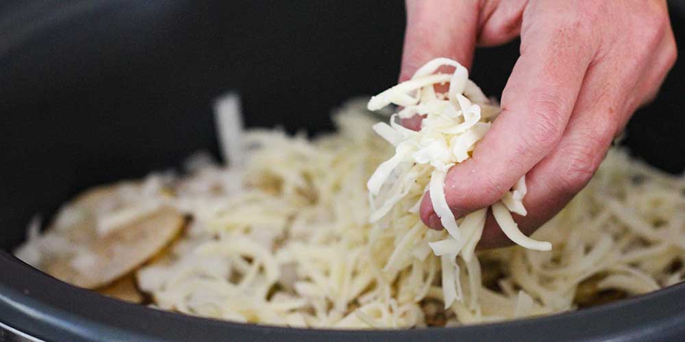 A hand dropping shredded Swiss cheese into a slow cooker lined with sliced potatoes.