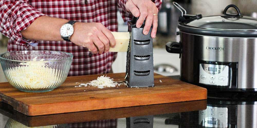 A hand grating a block of Swiss cheese on a box grater.