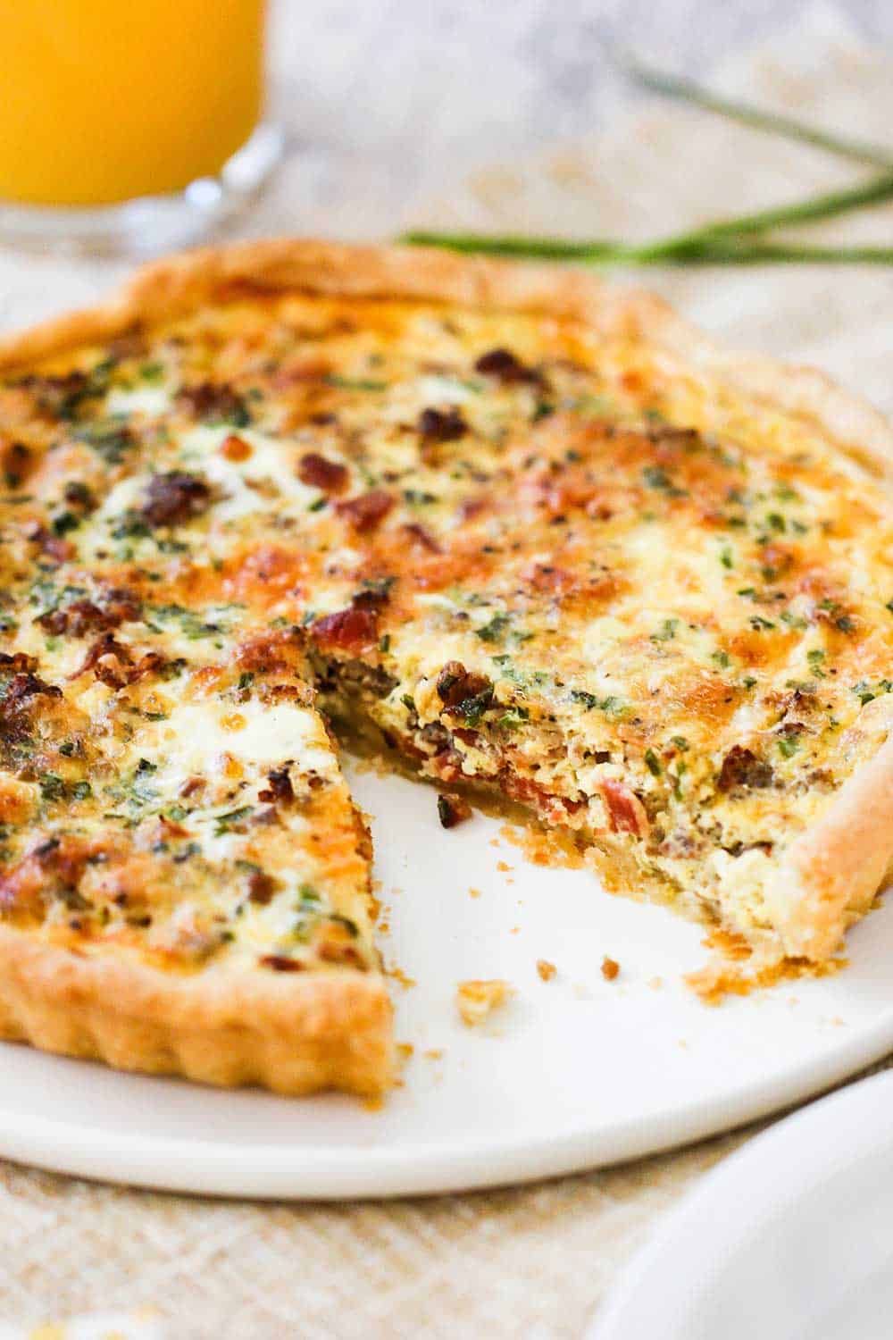 A breakfast quiche on a circular white platter with a slice missing and a glass of orange juice next to it.