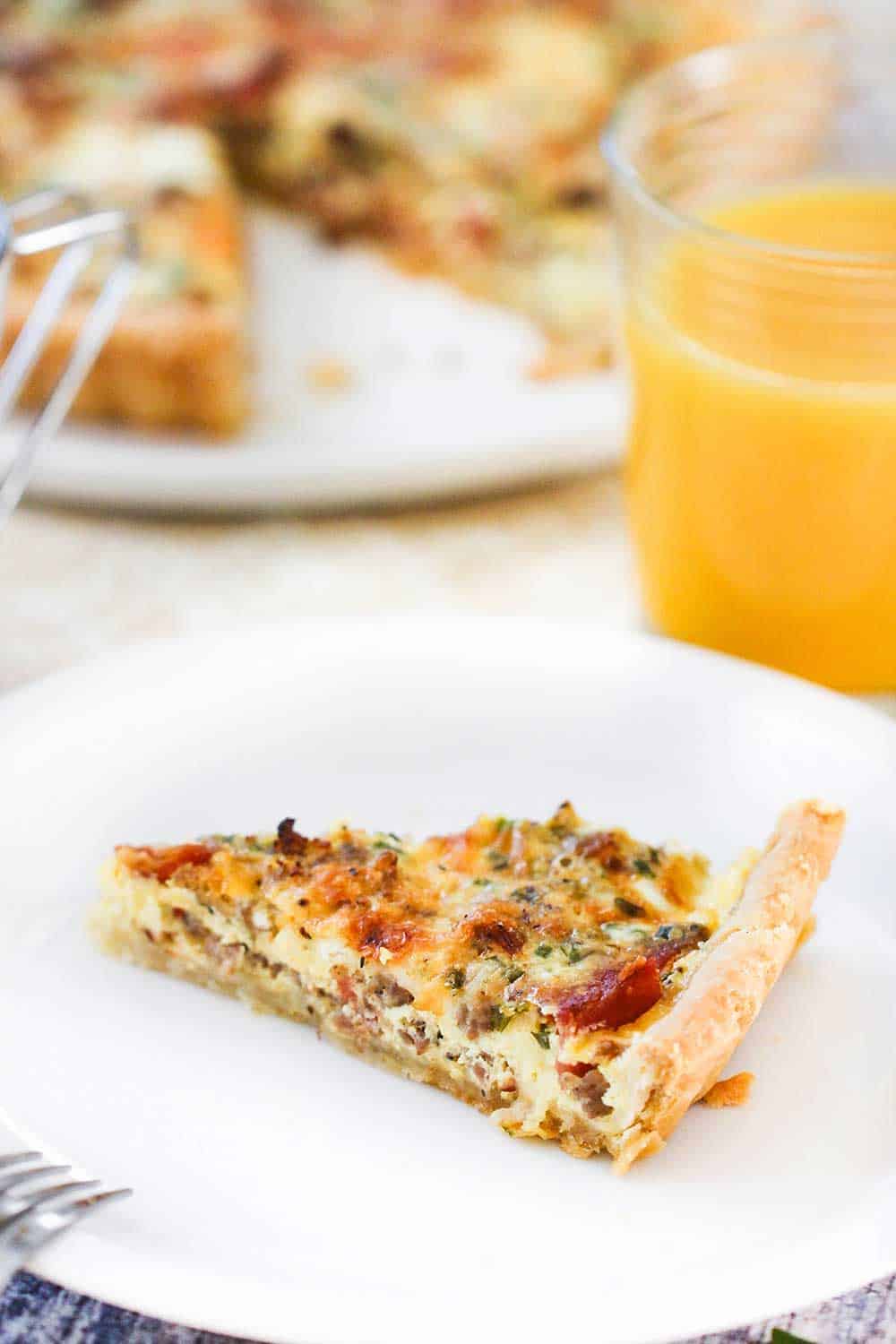 A slice of breakfast quiche on a white plate next to a glass of orange juice.