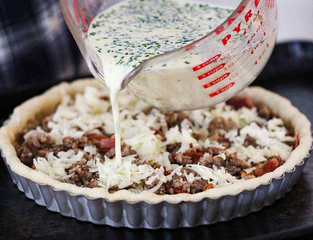 A large measuring cup pouring a cream mixture over sausage and cheese in a tart pan.