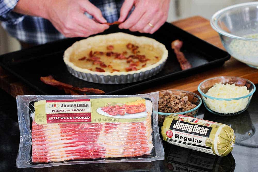 Two hands crumbling bacon into a tart pan lined with dough with bowls of cooked sausage nearby.