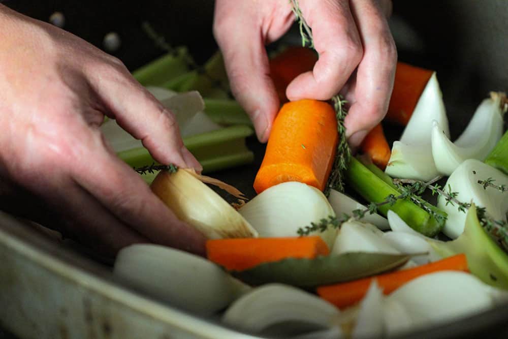 Two hands placing cut carrots, celery and onion in a large roasting pan.