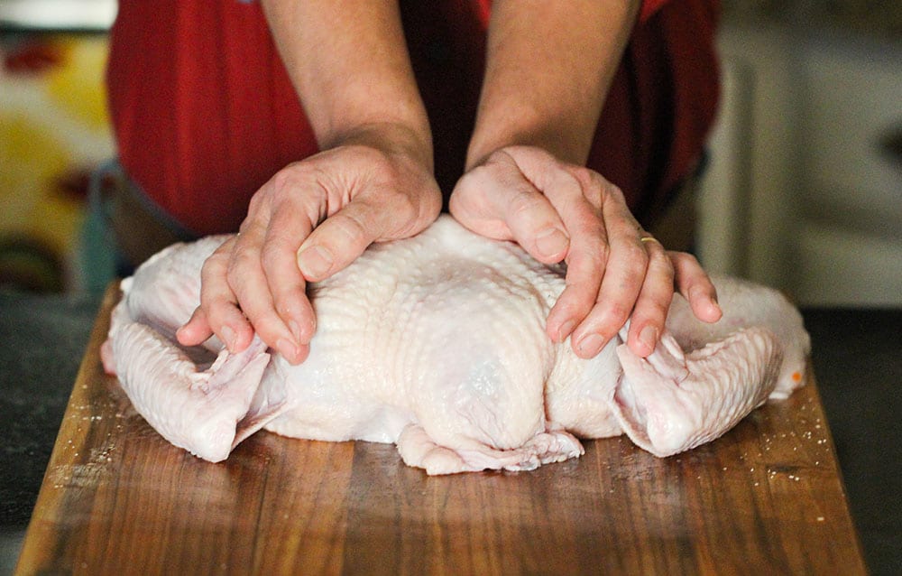 Two hands pressing down on an uncooked turkey that has been spatchcocked.