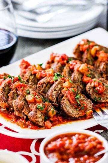 A white platter holding sliced slow cooker beef braciole with a bowl of marinara next to it.