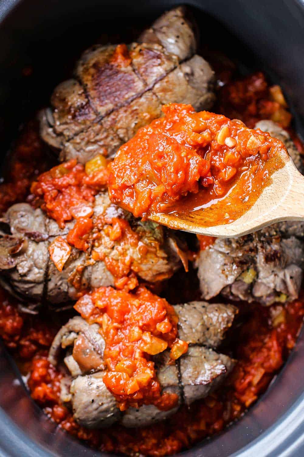 A wooden spoon adding marinara sauce onto rolls of beef in a slow cooker.