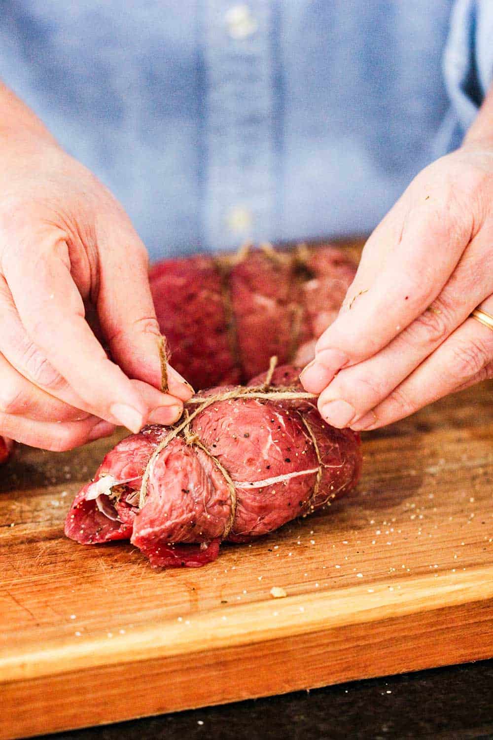 Two hands tying twine around a rolled up, stuffed flank steak.