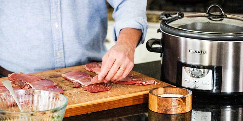 A hand laying a piece of prosciutto on a thin slice of round steak next to a slow cooker.