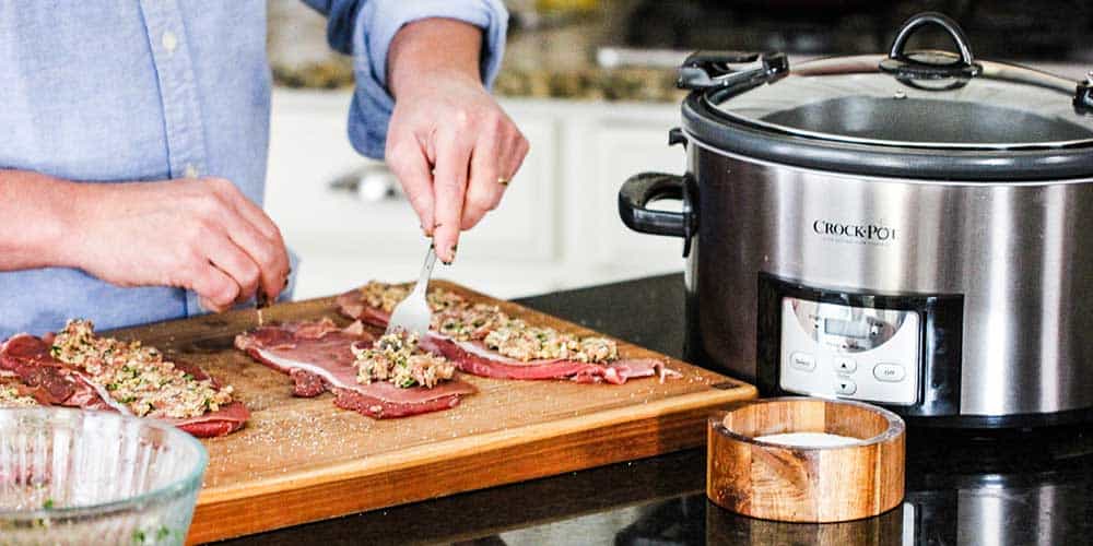 A hand using a fork to spread filling onto a piece of steak for beef braciole.