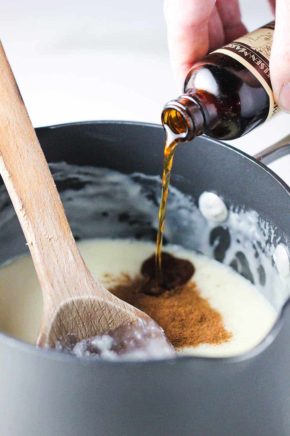 A hand pouring vanilla extract from a bottle into a pan of snickerdoodle fudge.