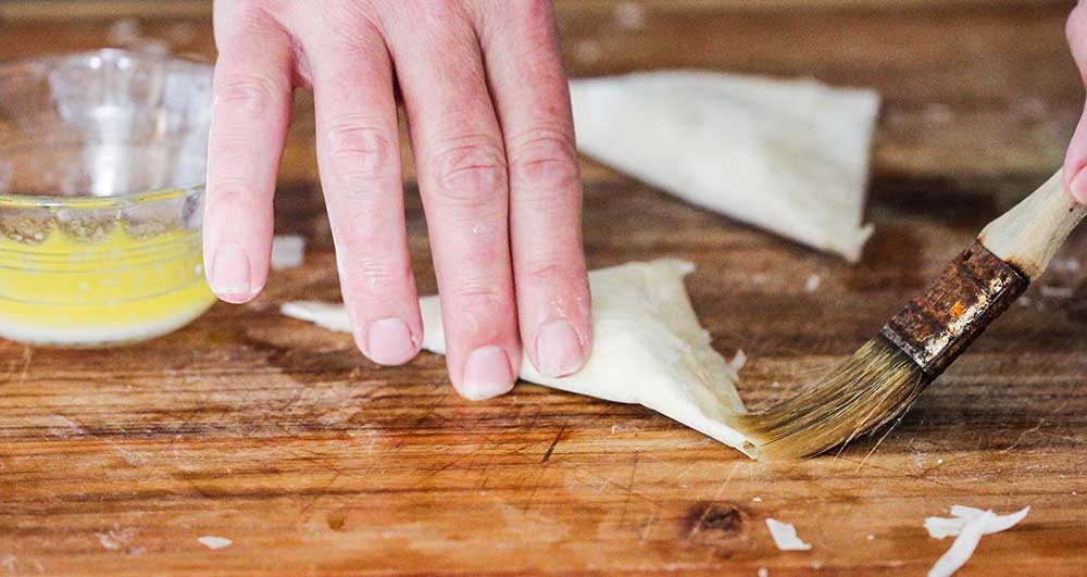 Two hands applying butter with a brush to an air fryer shrimp and artichoke phyllo triangle.