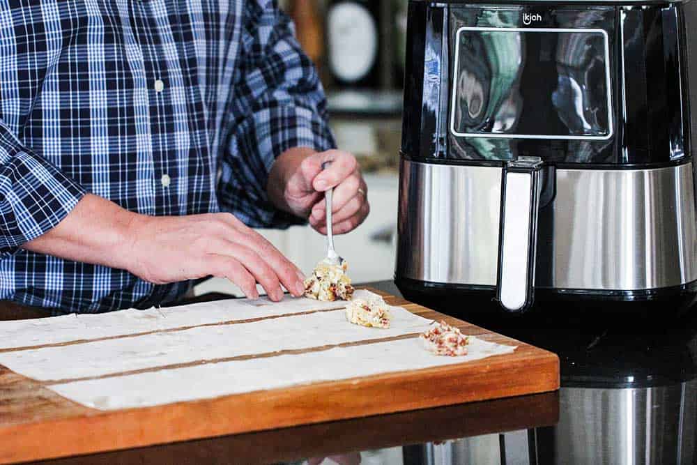 Two hands spooning a shrimp and artichoke mixture on to the ends of phyllo dough strips next to an air fryer.