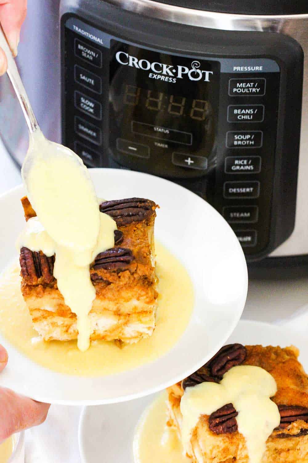 A spoon pouring custard onto a plate of pumpkin bread pudding next to a pressure cooker.