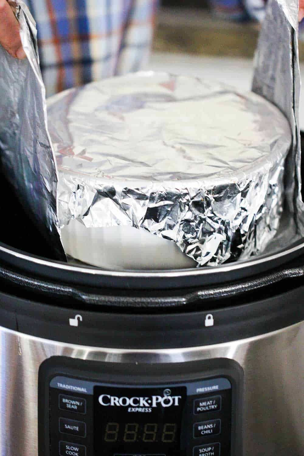 A dish covered in foil being lowered by a foil sling into a pressure cooker.