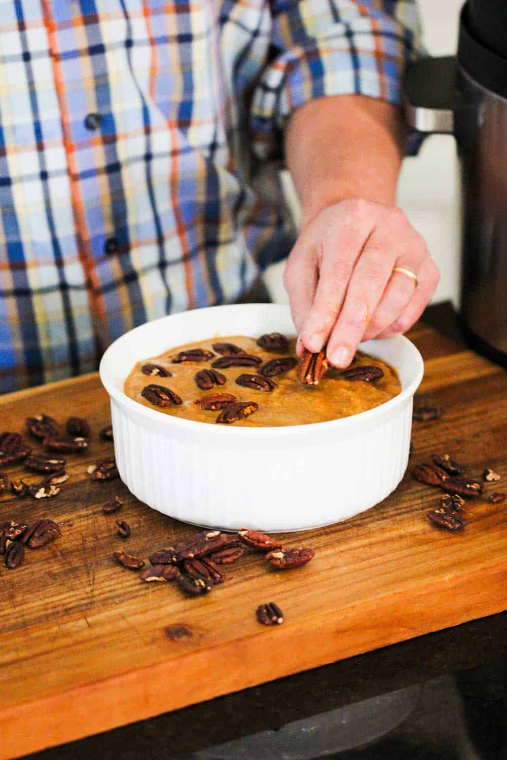 A hand placing pecans on top of a pumpkin bread pudding in a white bowl
