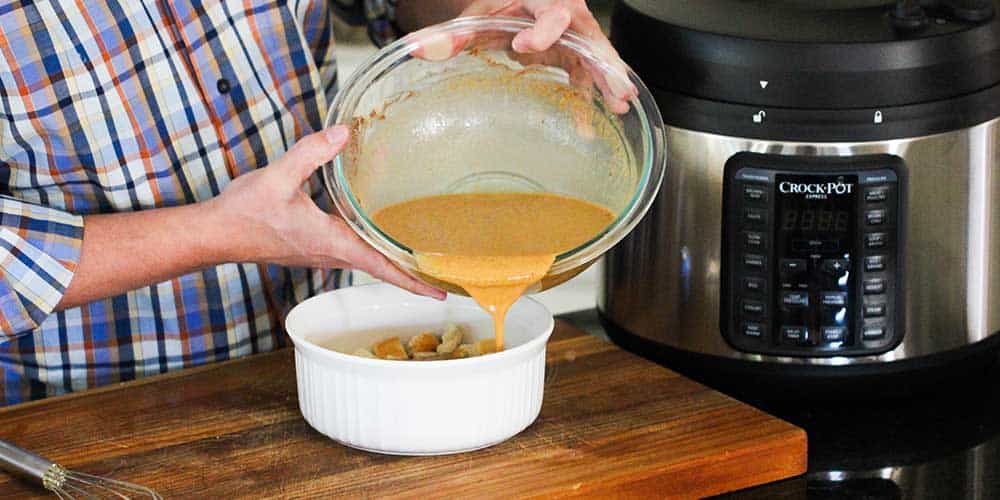 Two hands pouring a bowl of an egg mixture into a white bowl filled with bread chunks.