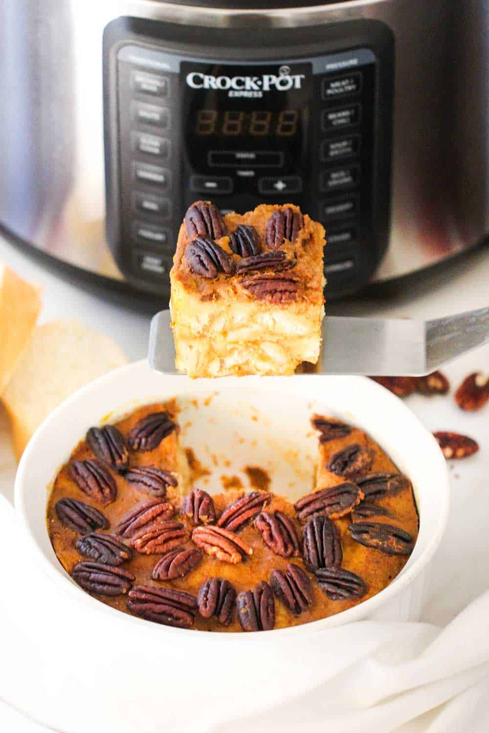 A slice of pumpkin bread pudding being held up on a spatula next to a Crock Pot Pressure Cooker.