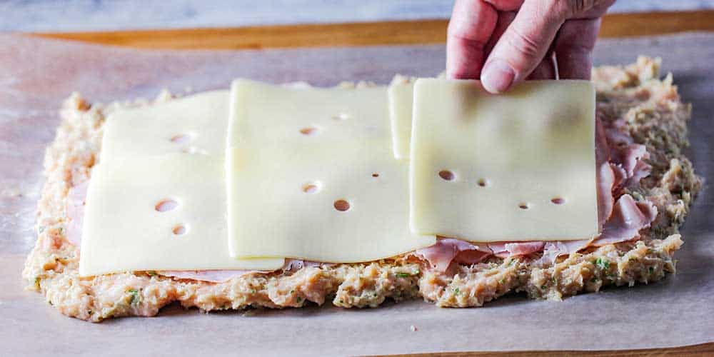 A hand placing sliced of Swiss cheese on ham slices on a layer of chicken meatloaf on wax paper.