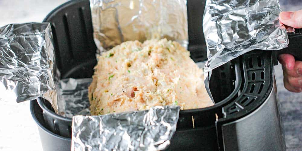 A chicken cordon bleu loaf being lowered into an air fryer with two aluminum foil slings.