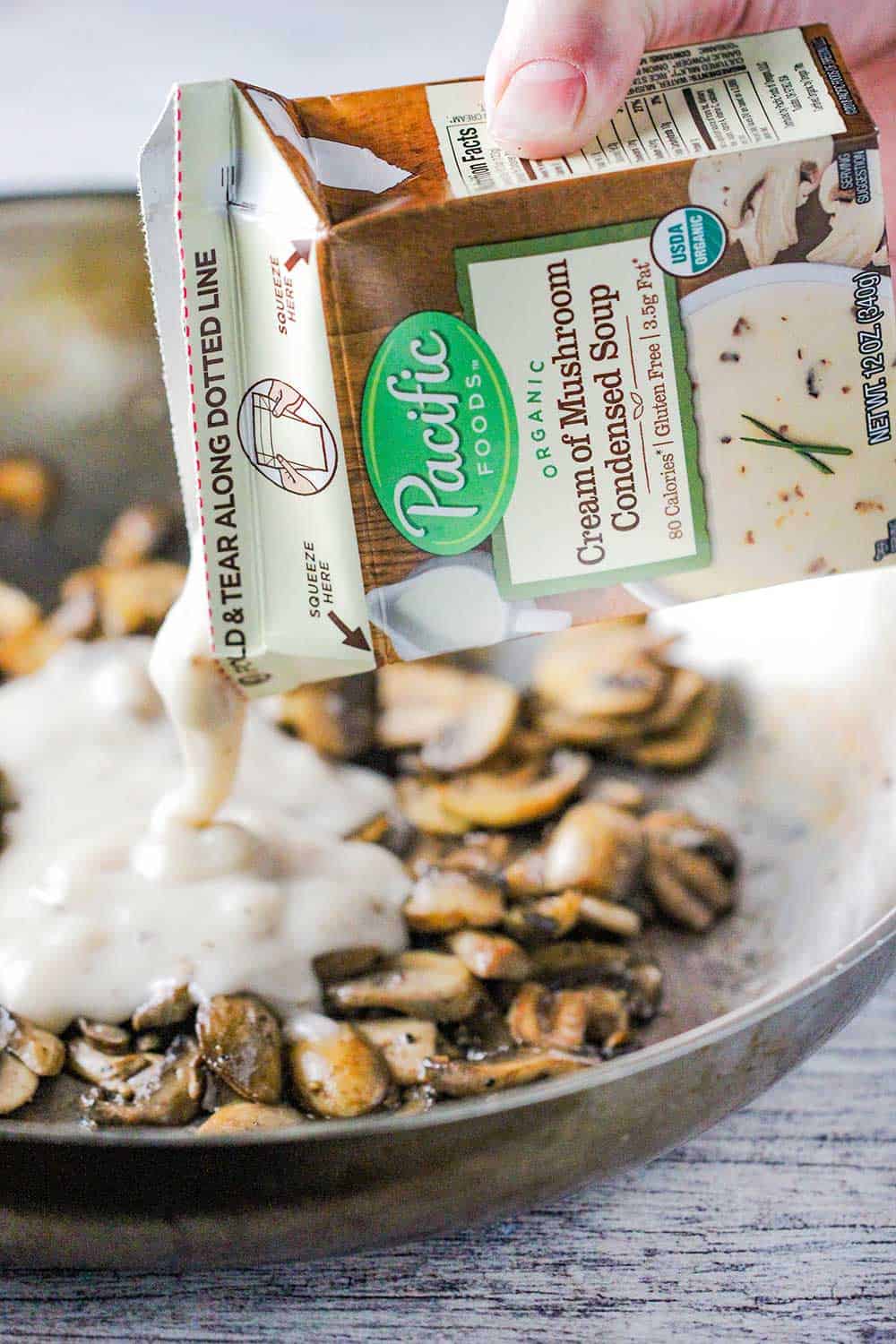 A hand pouring a box of condensed cream of mushroom soup in a skillet of sautéed mushrooms.